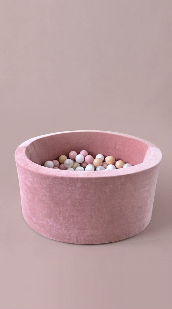 Rond roze ballenbad met fluweelachtige bekleding en witte, roze en beige ballen tegen een roze achtergrond
