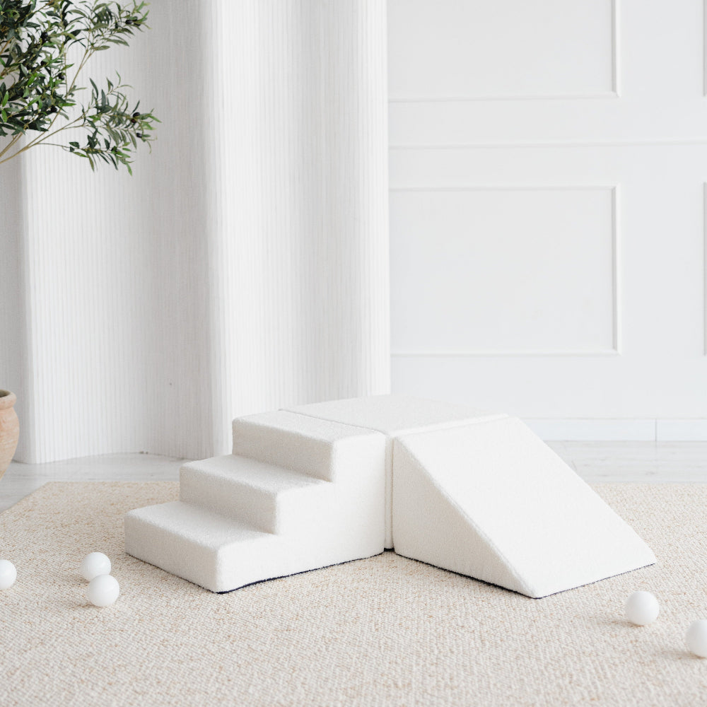 Witte driedelige bouclé speelset met trapje, blok en driehoek op een beige vloerkleed, met witte ballen eromheen.
