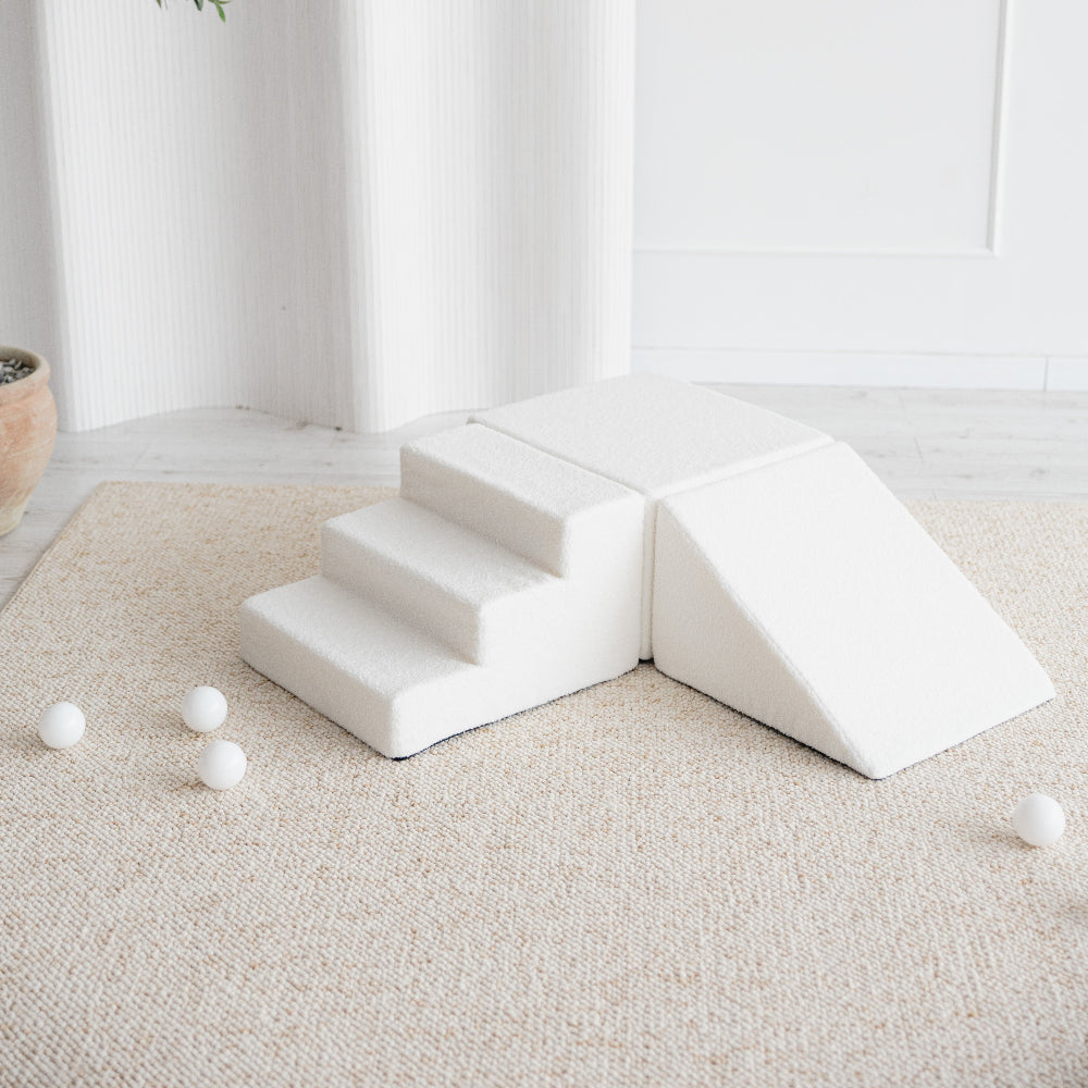 Witte bouclé speelset met trapje, blok en driehoekige helling, schuin van boven gezien op een beige vloerkleed.