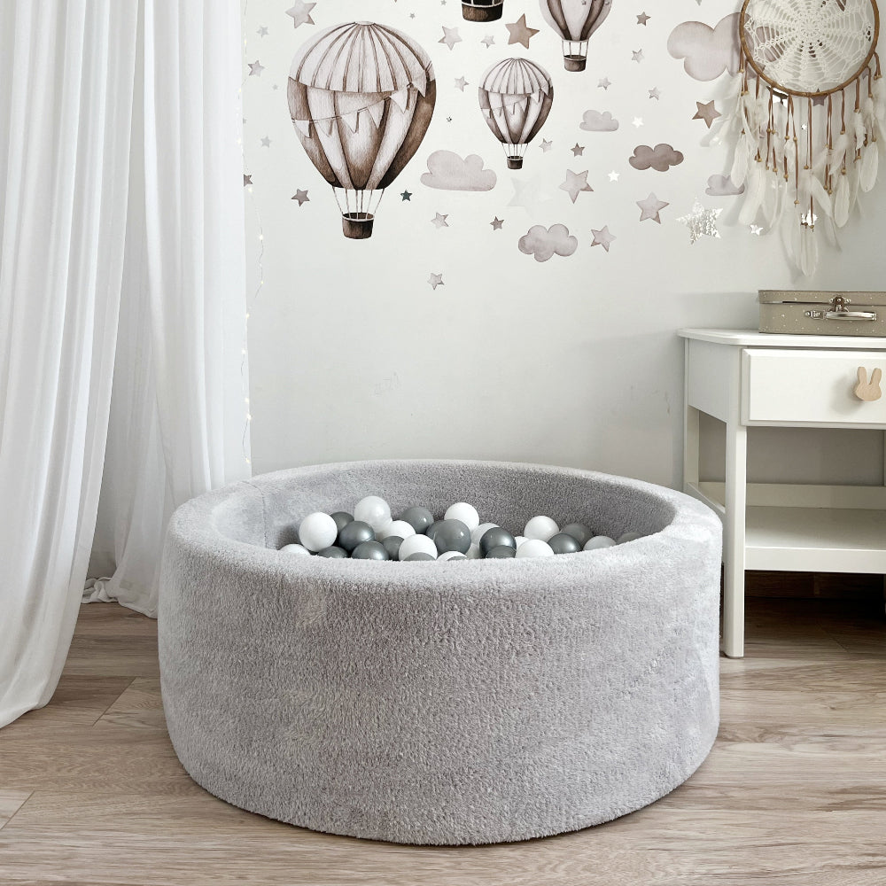 Rond lichtgrijs teddy ballenbad met witte en grijze balletjes in een kinderkamer met luchtballonnen op de muur.