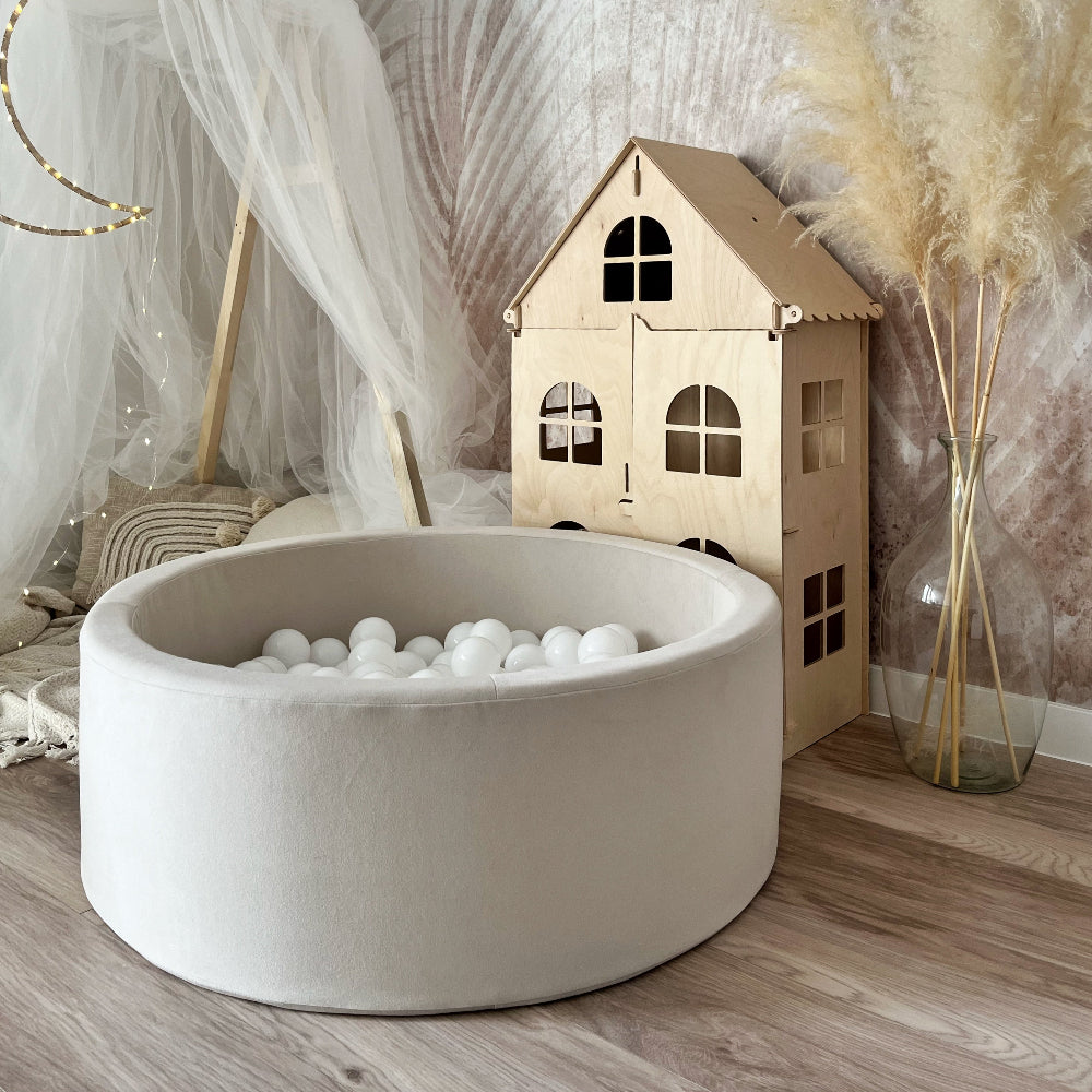 Rond crèmekleurig ballenbad met witte balletjes naast een houten poppenhuis en een witte klamboe.