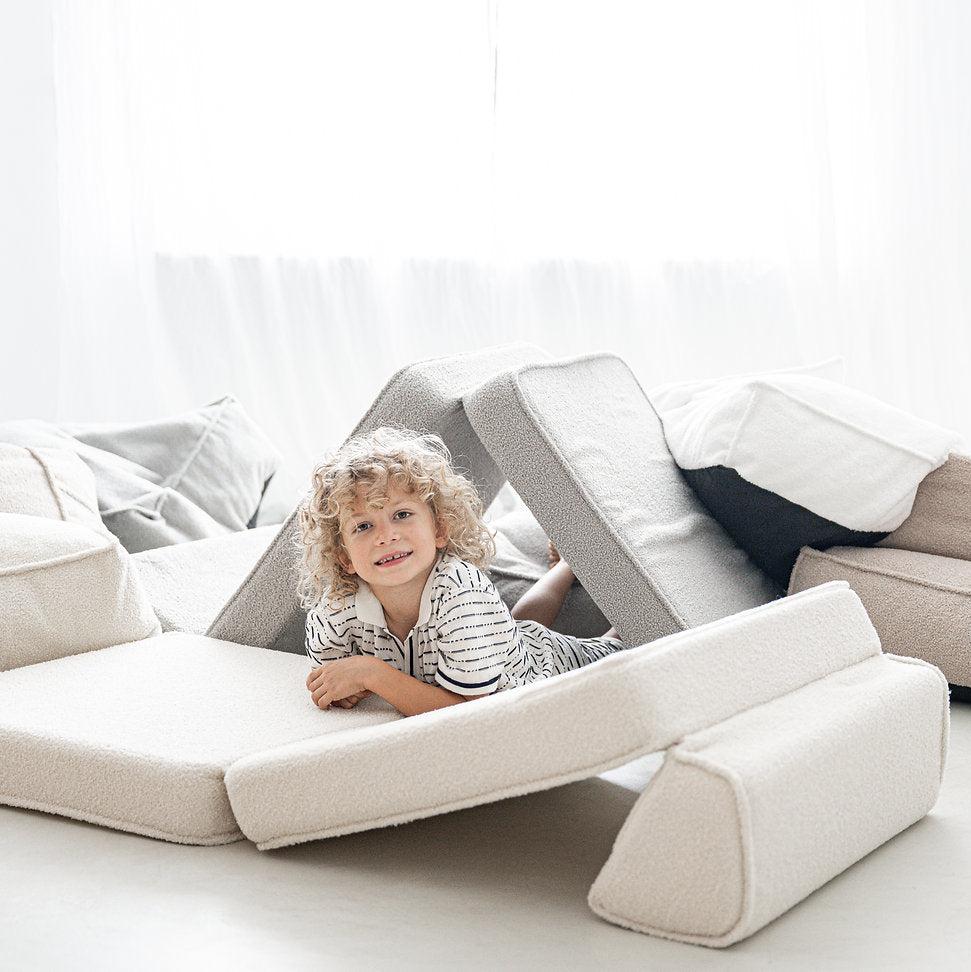 Kind ligt onder een driehoek van loungersegmenten, met witte bouclé kussens op de voorgrond.