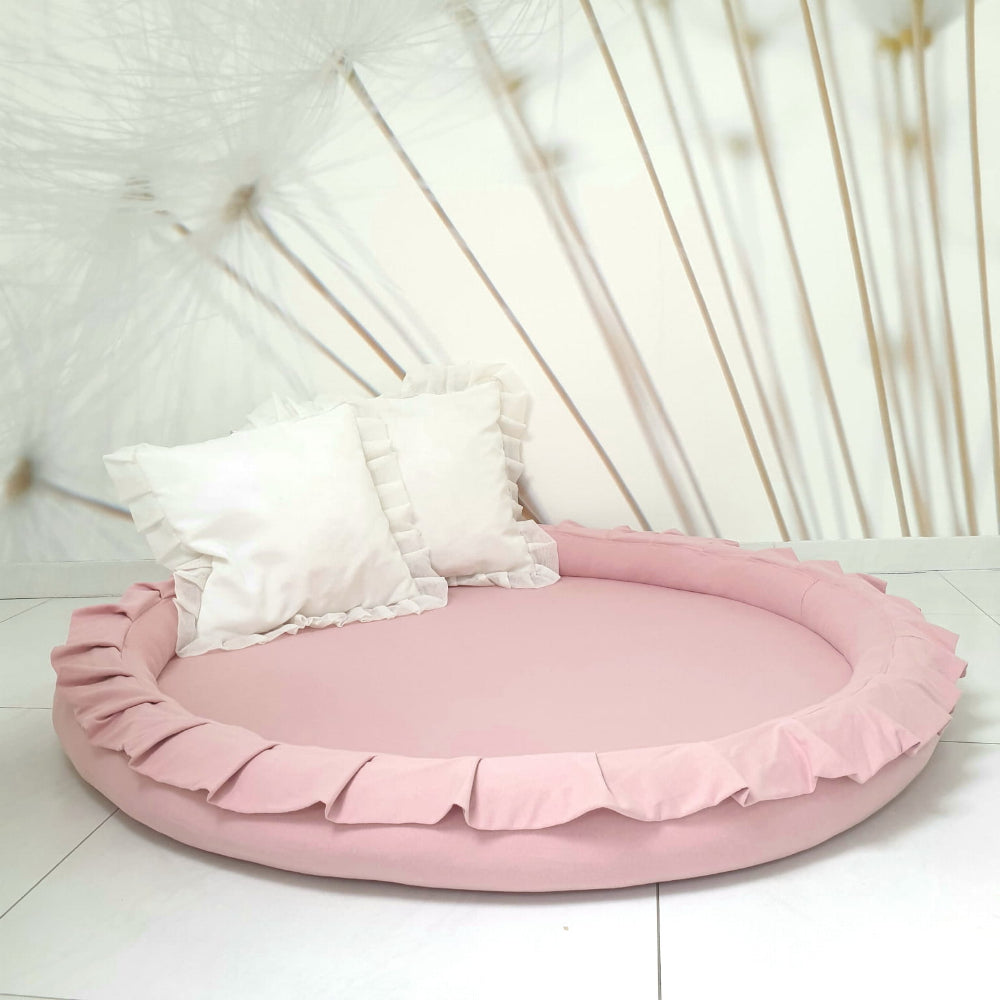 Rond roze speelnest met opstaande rand en ruches, met twee witte kussens op een lichte tegelvloer.